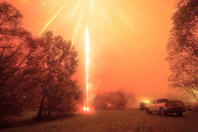 A firework lights up the night sky, illuminating trees and a pickup truck.