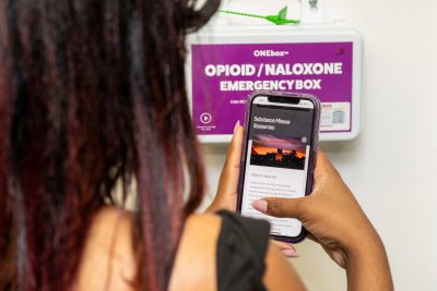 A ONEbox opioid overdose reversal kit on a wall and a person viewing its video instructions on their phone, seen over their shoulder.